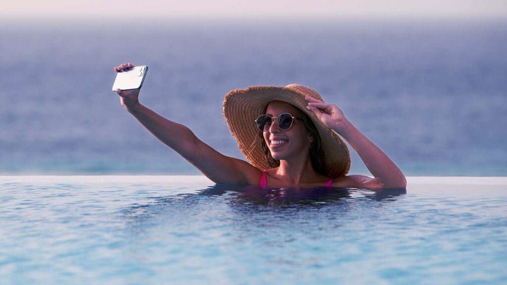Beuatiful woman with a hat, inside the infinity swimming pool overlooking the ocean taking a selfie herself with her left hand touching the brown hat.