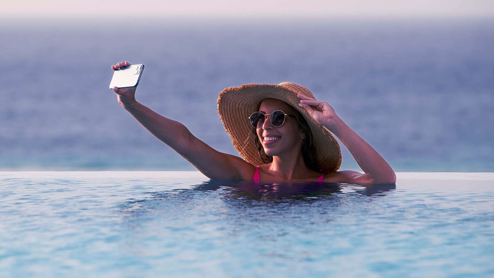 Beuatiful woman with a hat, inside the infinity swimming pool overlooking the ocean taking a selfie herself with her left hand touching the brown hat.